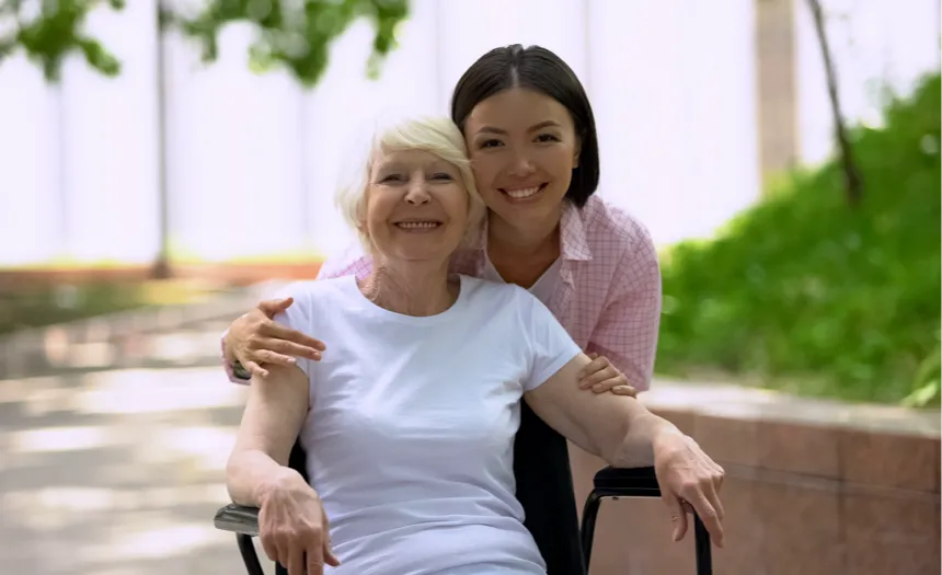 A happy, elderly woman in a wheelchair being embraced from behind by a younger woman, possibly her caregiver or family member. Both women are smiling warmly, with the elderly woman wearing a white shirt and the younger woman dressed in a pink plaid shirt. They are outdoors in a peaceful, leafy setting with soft sunlight filtering through the trees in the background, giving the scene a sense of warmth and connection.